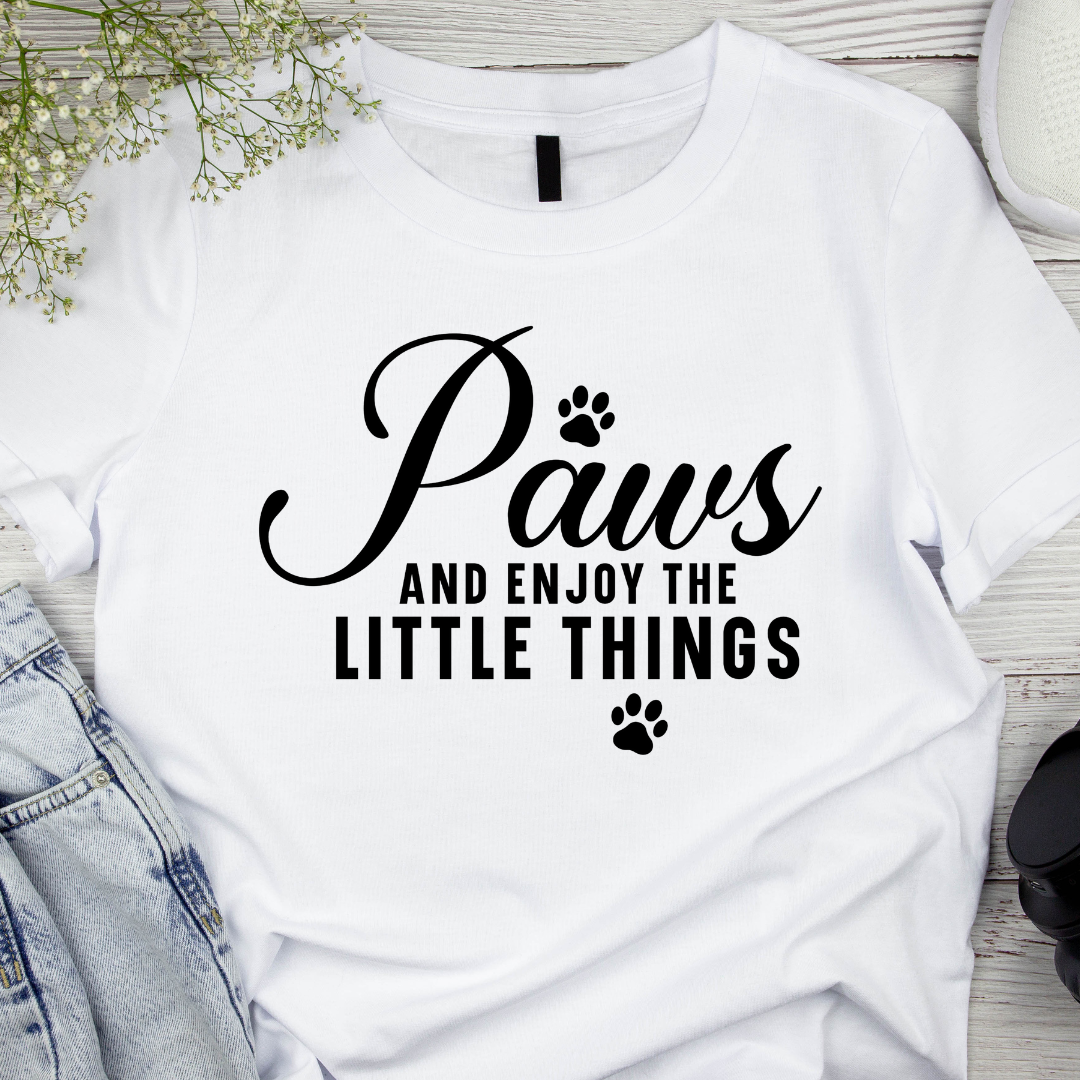 White t-shirt with 'Paws and enjoy the little things' text on a wooden surface.