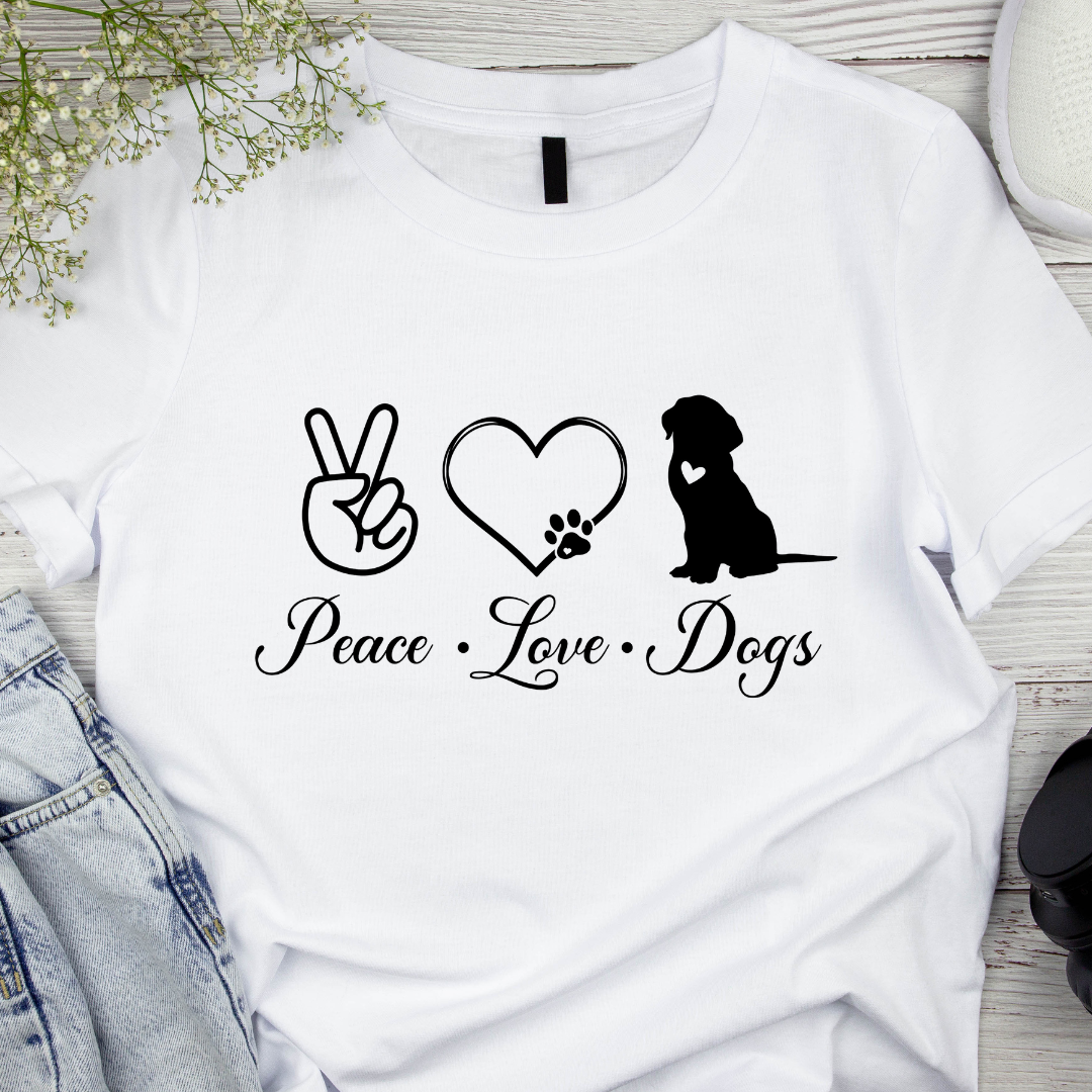 White t-shirt with 'Peace Love Dogs' design featuring a peace sign, heart with paw print, and dog silhouette on a wooden surface.
