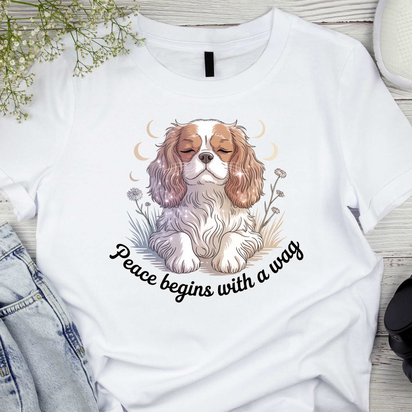 White t-shirt with a dog illustration and 'Peace begins with a wag' text on a wooden surface.