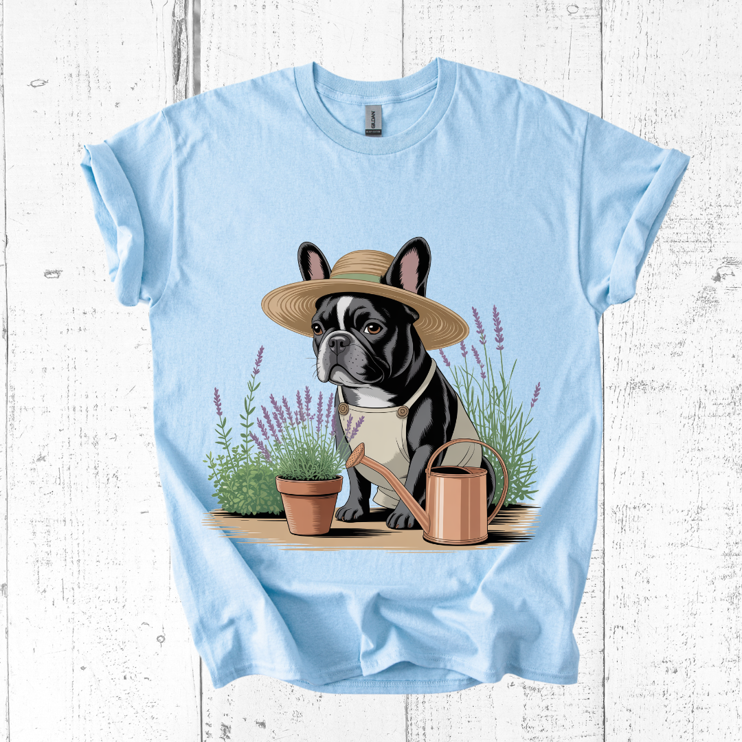 Light blue t-shirt with a graphic of a dog wearing a straw hat and holding a watering can, standing on a wooden platform with plants and lavender flowers.