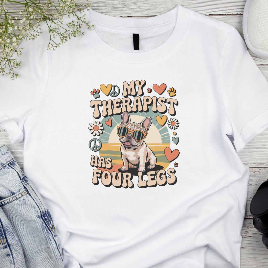 White t-shirt with a graphic of a dog wearing sunglasses and text about a therapist with four legs, on a wooden surface.