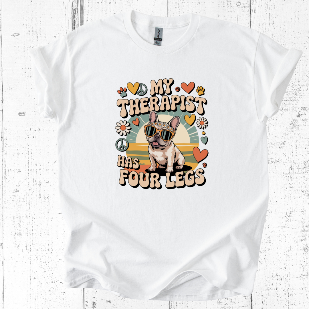 White t-shirt with a dog graphic and text 'My therapist has four legs' on a wooden surface.