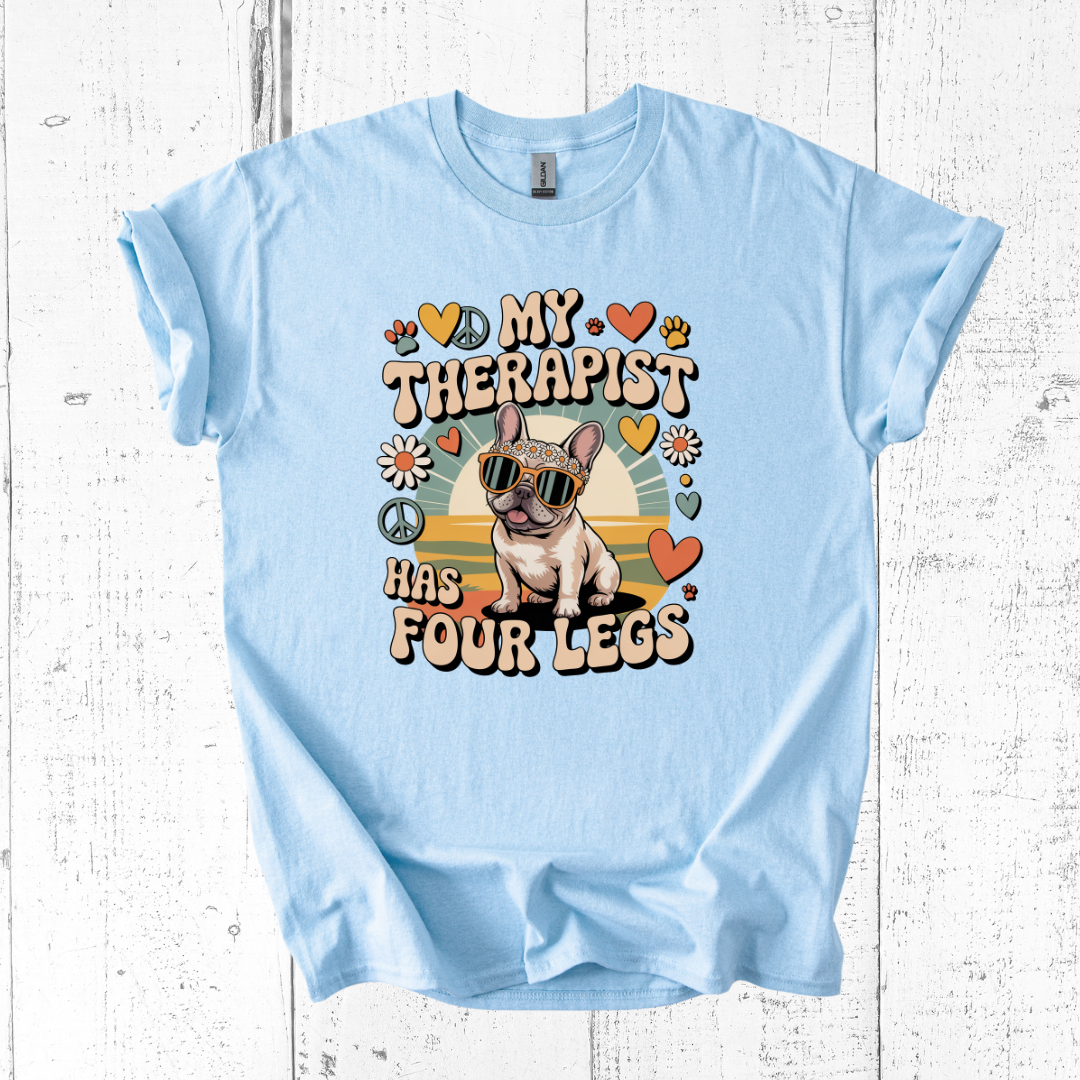 Light blue t-shirt with a graphic of a dog and text 'My therapist has four legs' on a white wooden background.