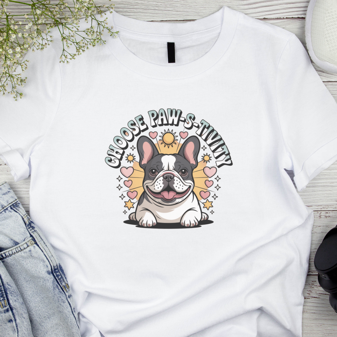White t-shirt with a cartoon bulldog and 'Choose PAW-sitivity' text on a wooden surface.