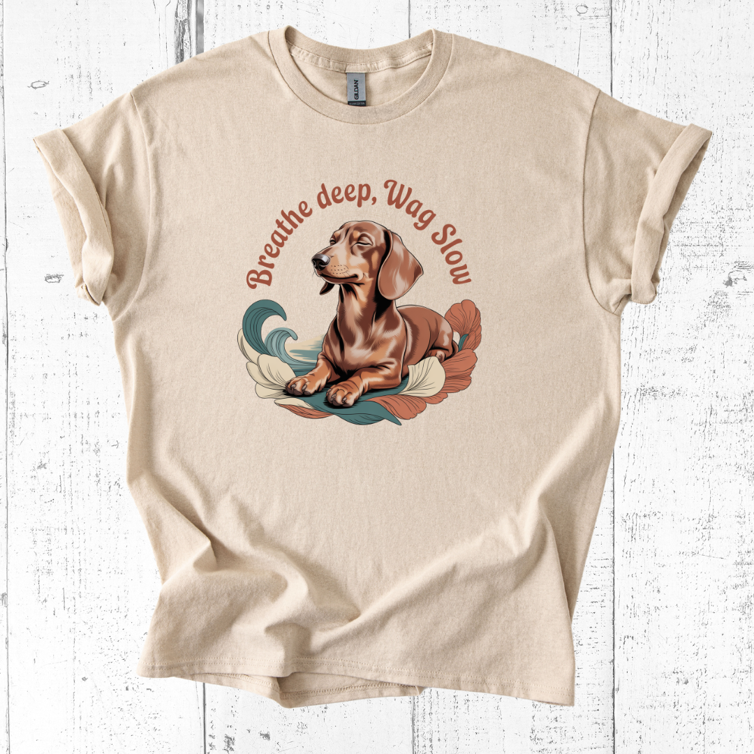 Beige t-shirt with a dachshund graphic and text 'Breathe deep, Wag Slow' on a wooden surface.