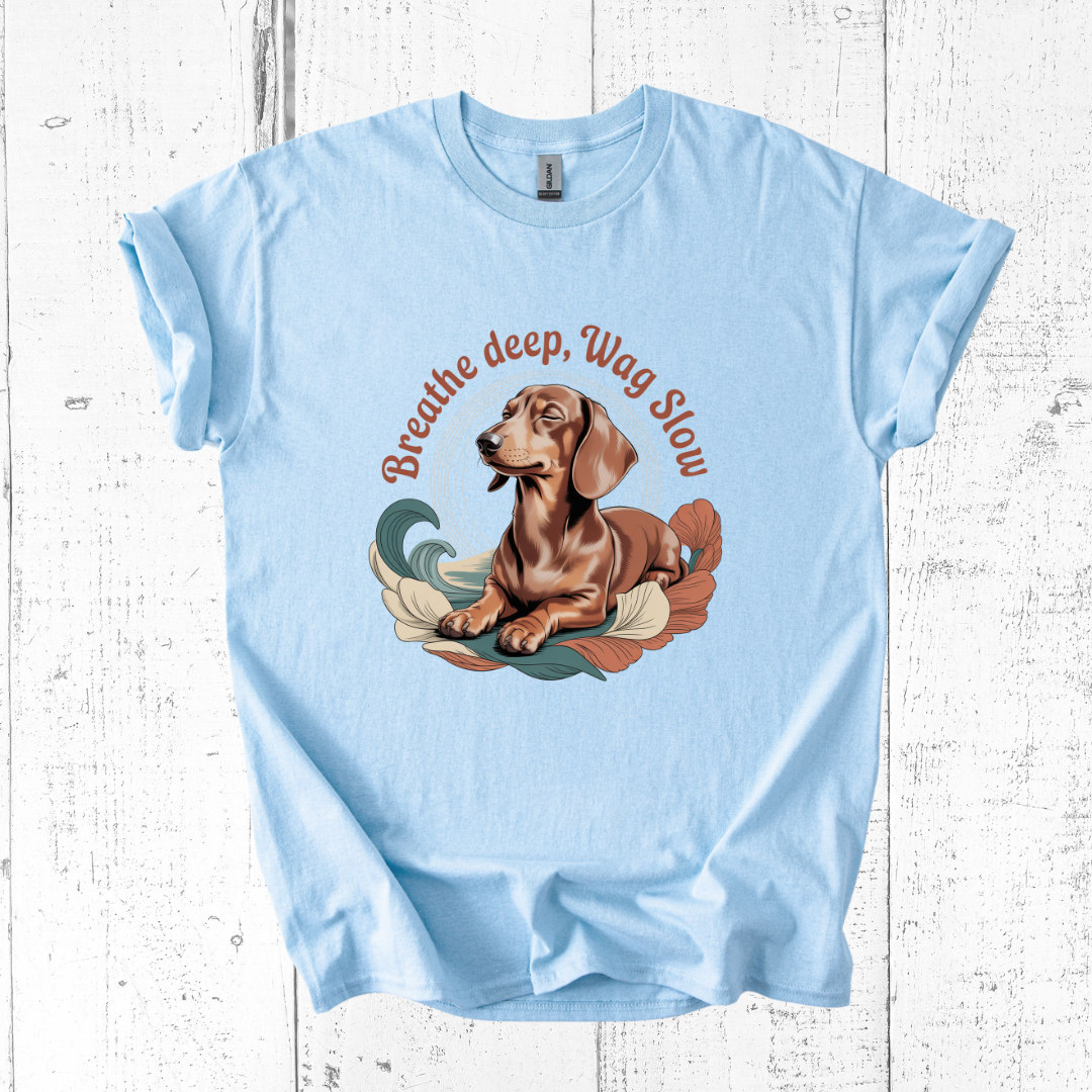 Light blue t-shirt with a dachshund graphic and text 'Breathe deep, Wag Slow' on a wooden surface.