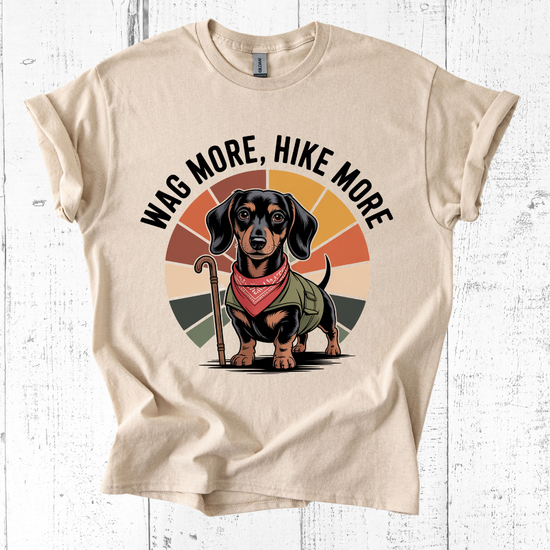 Beige t-shirt with a dachshund graphic and 'Wag More, Hike More' text on a white wooden background