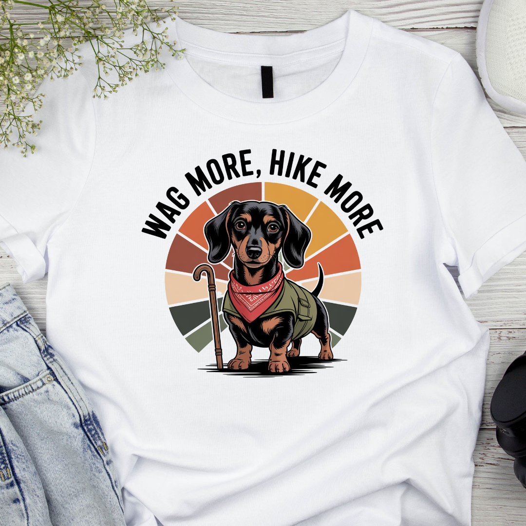 White t-shirt with a dachshund graphic and 'Wag More, Hike More' text on a wooden surface.
