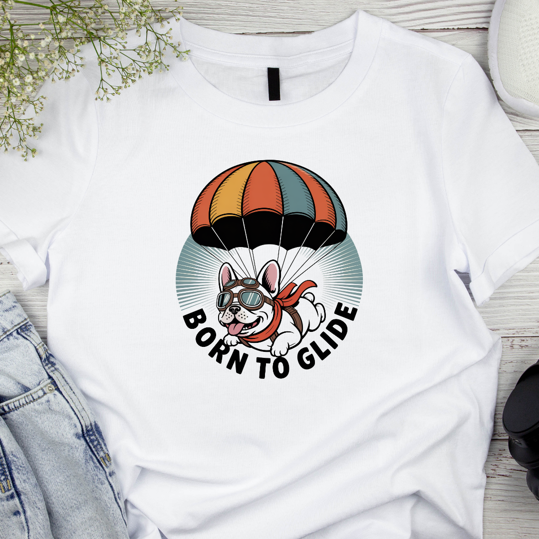 White t-shirt with a cartoon dog paragliding design and 'Born to Glide' text on a wooden surface.
