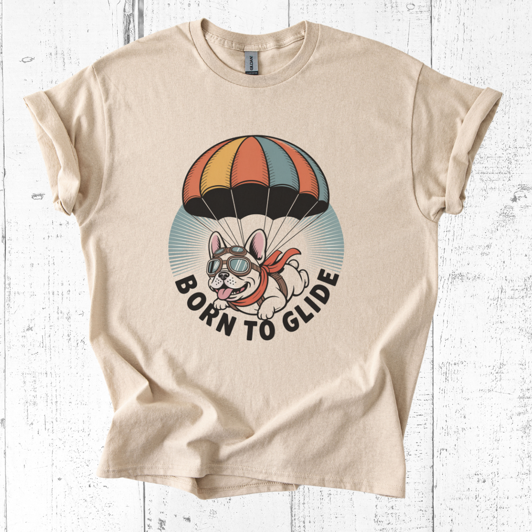 Beige t-shirt with a graphic of a dog parachuting and 'Born to Glide' text on a white wooden background.