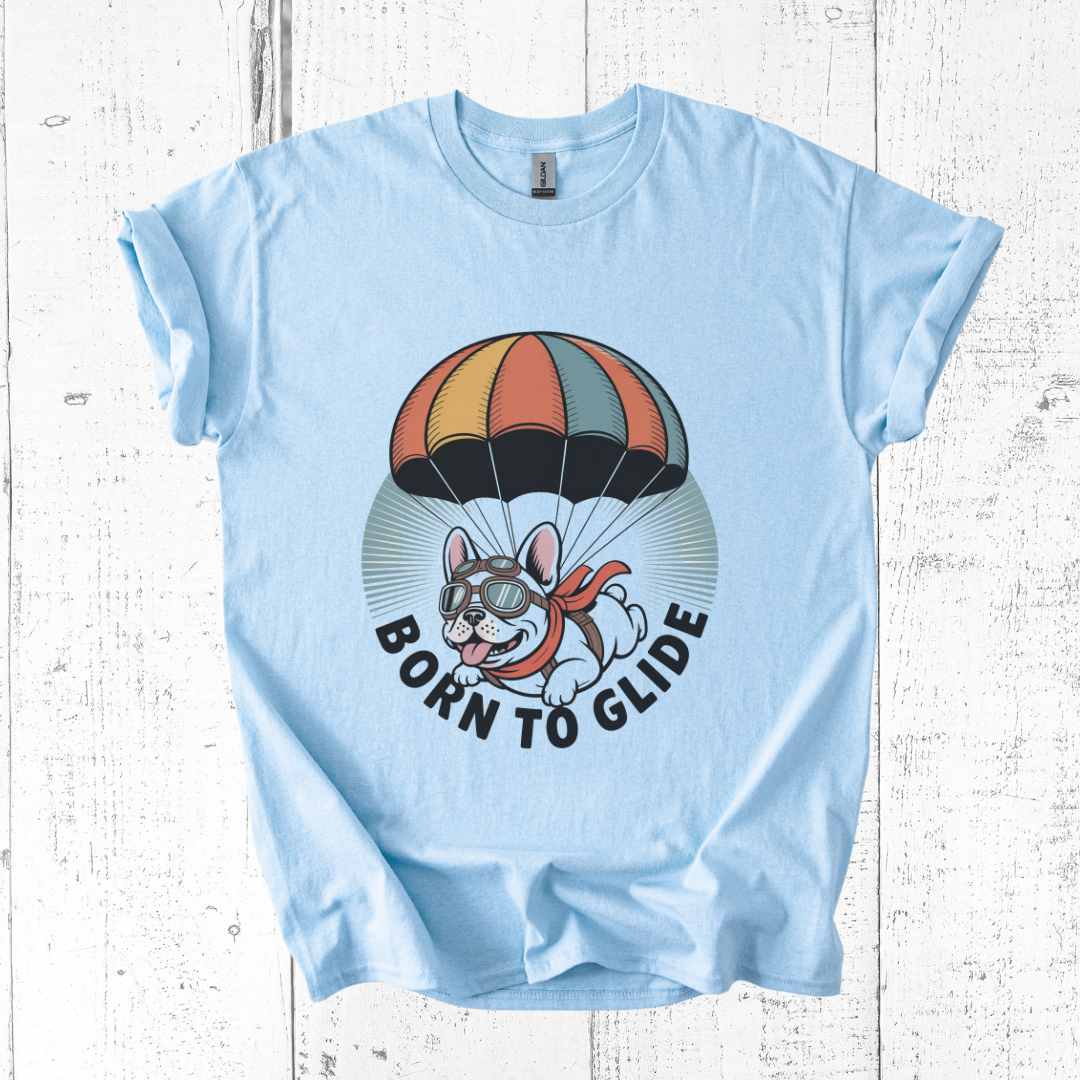 Light blue t-shirt with a graphic of a dog with goggles and a parachute, and the text 'Born to Glide' on a white wooden background.