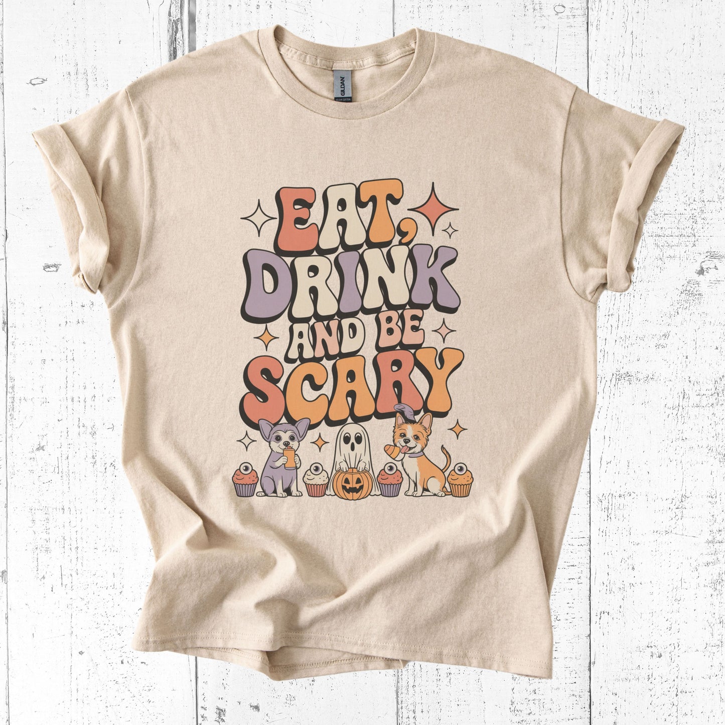 Beige t-shirt with Halloween-themed graphic and text 'Eat, Drink and Be Scary' on a white wooden background.