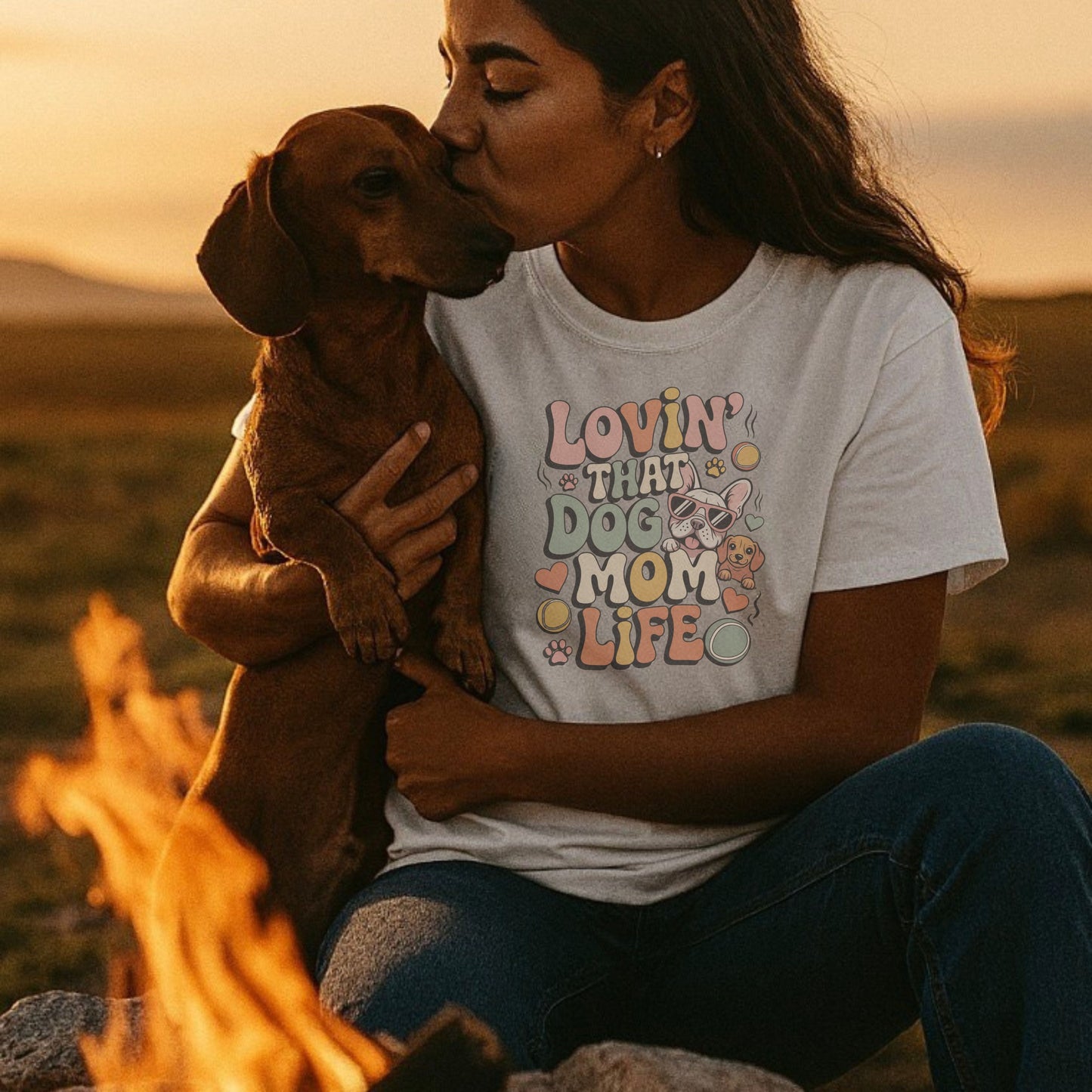 Woman holding a dog and wearing a 'Lovin' That Dog Mom Life' shirt by a campfire.