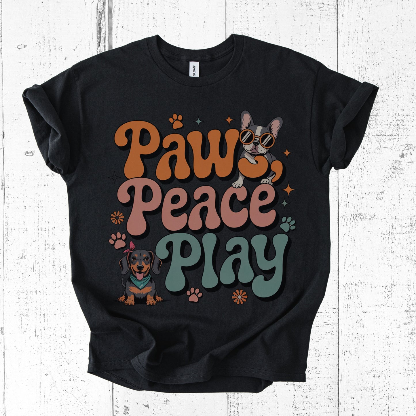 Black t-shirt with 'Paws Peace Play' text and dog illustrations on a white wooden background