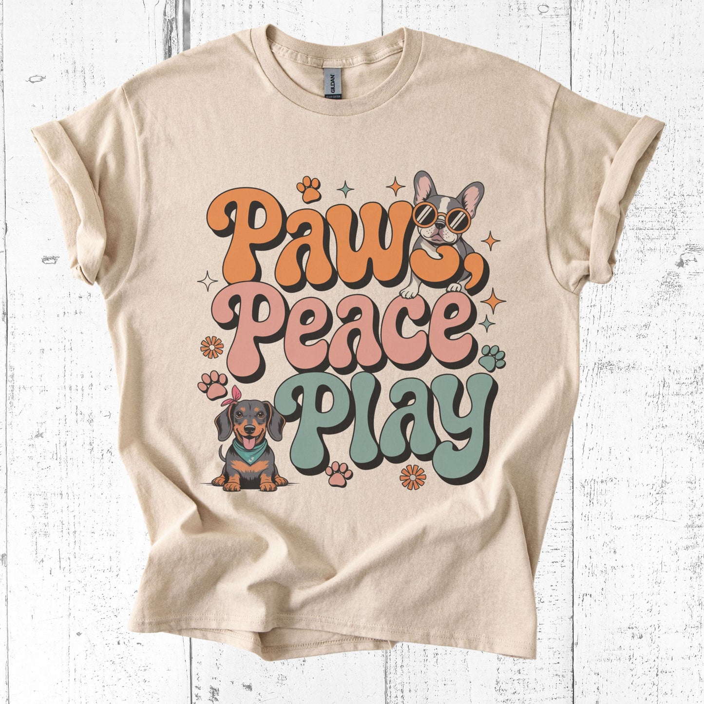 Beige t-shirt with 'Paws Peace Play' text and dog illustrations on a white wooden surface