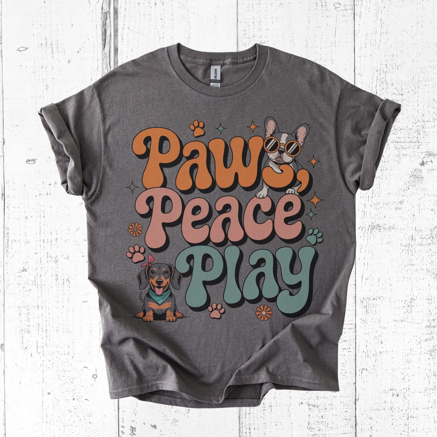 Gray t-shirt with 'Paws Peace Play' text and dog illustrations on a white wooden surface