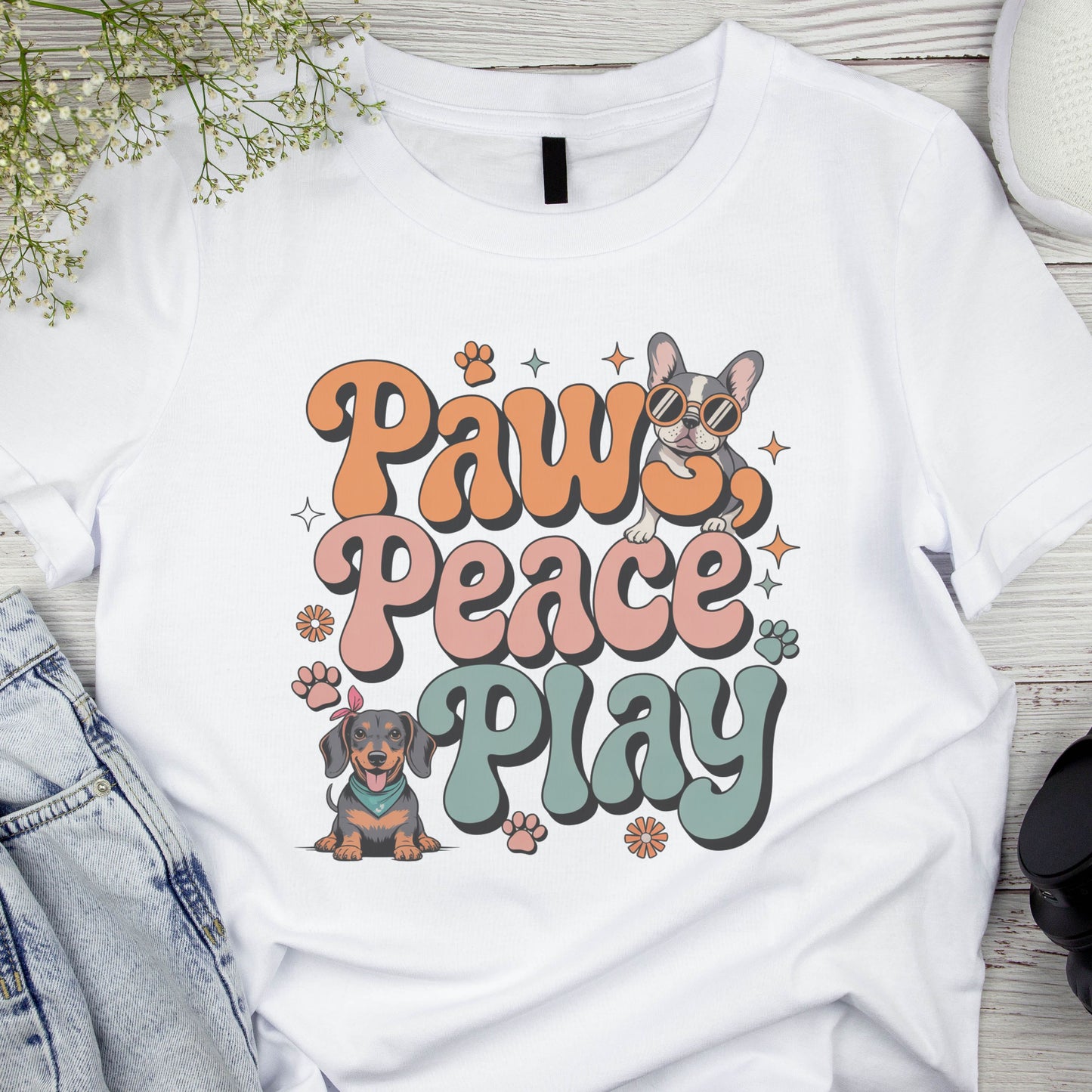 White t-shirt with 'Paw Peace Play' text and dog illustrations on a wooden surface.