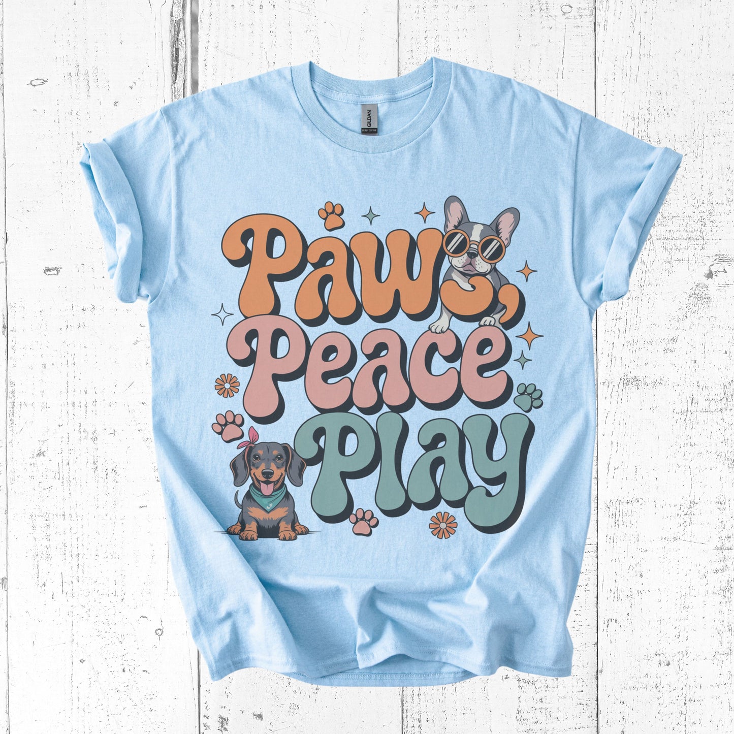 Light blue t-shirt with 'Paws Peace Play' text and dog illustrations on a wooden surface