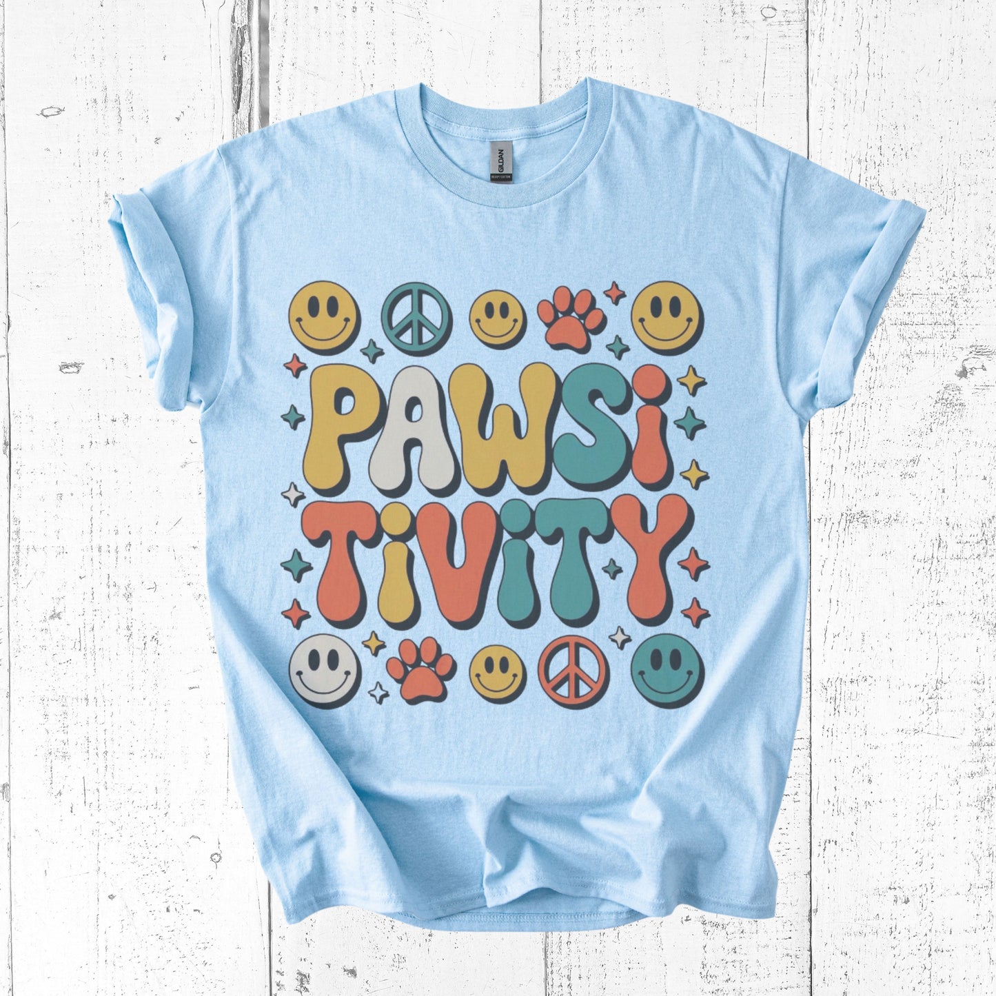 Light blue t-shirt with colorful 'PAWSI TIVITY' design on a white wooden background