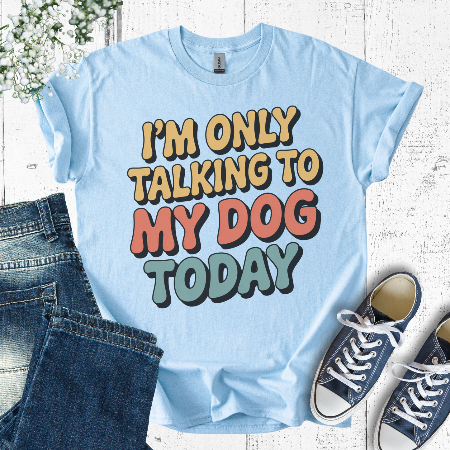 Light blue t-shirt with colorful text 'I'm only talking to my dog today' on a wooden surface with jeans and sneakers.