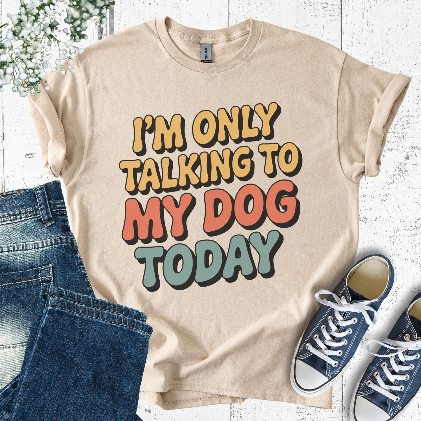 Beige t-shirt with colorful text 'I'm only talking to my dog today' on a white wooden surface with jeans and sneakers.