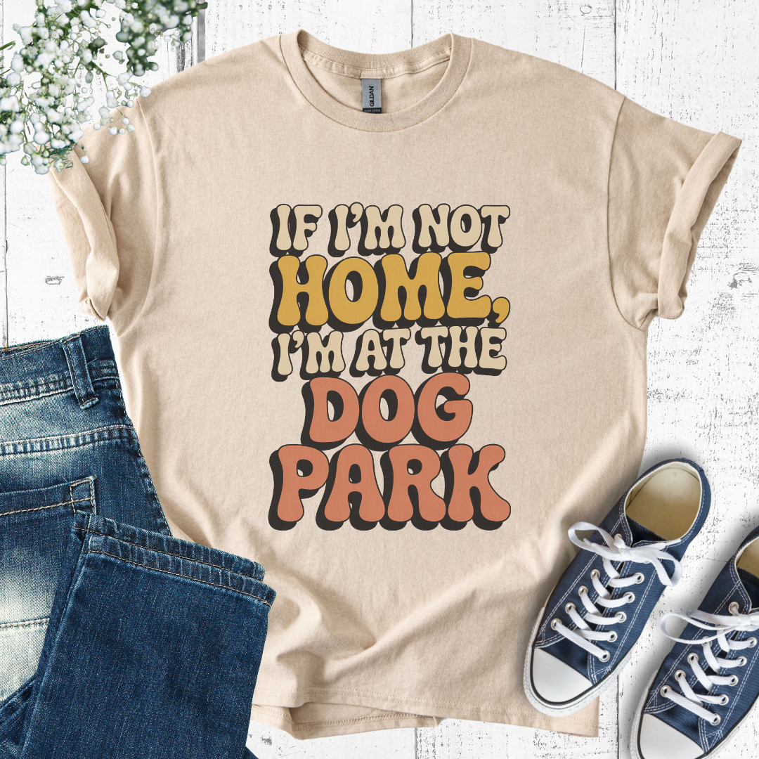 Beige t-shirt with 'If I'm not home, I'm at the dog park' text, paired with jeans and sneakers on a light wooden surface.