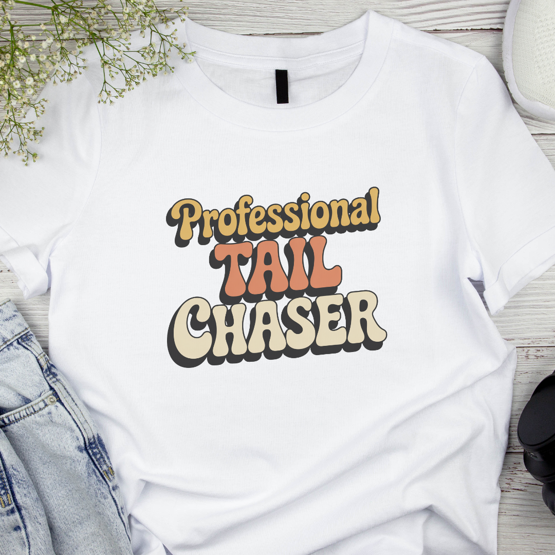 White t-shirt with 'Professional Tail Chaser' text on a wooden surface with denim jeans and flowers.