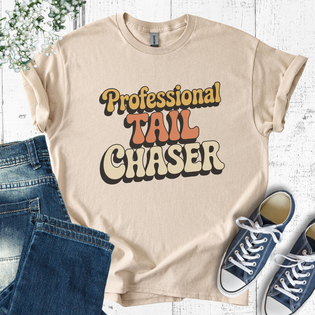 Beige t-shirt with 'Professional Tail Chaser' text, paired with jeans and sneakers on a light wooden surface.