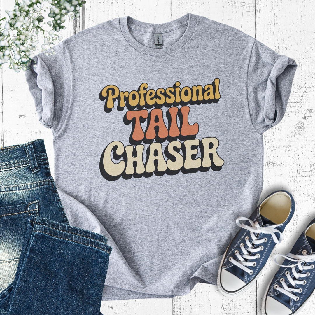 Gray t-shirt with 'Professional Tail Chaser' text, paired with jeans and sneakers on a light wooden surface.