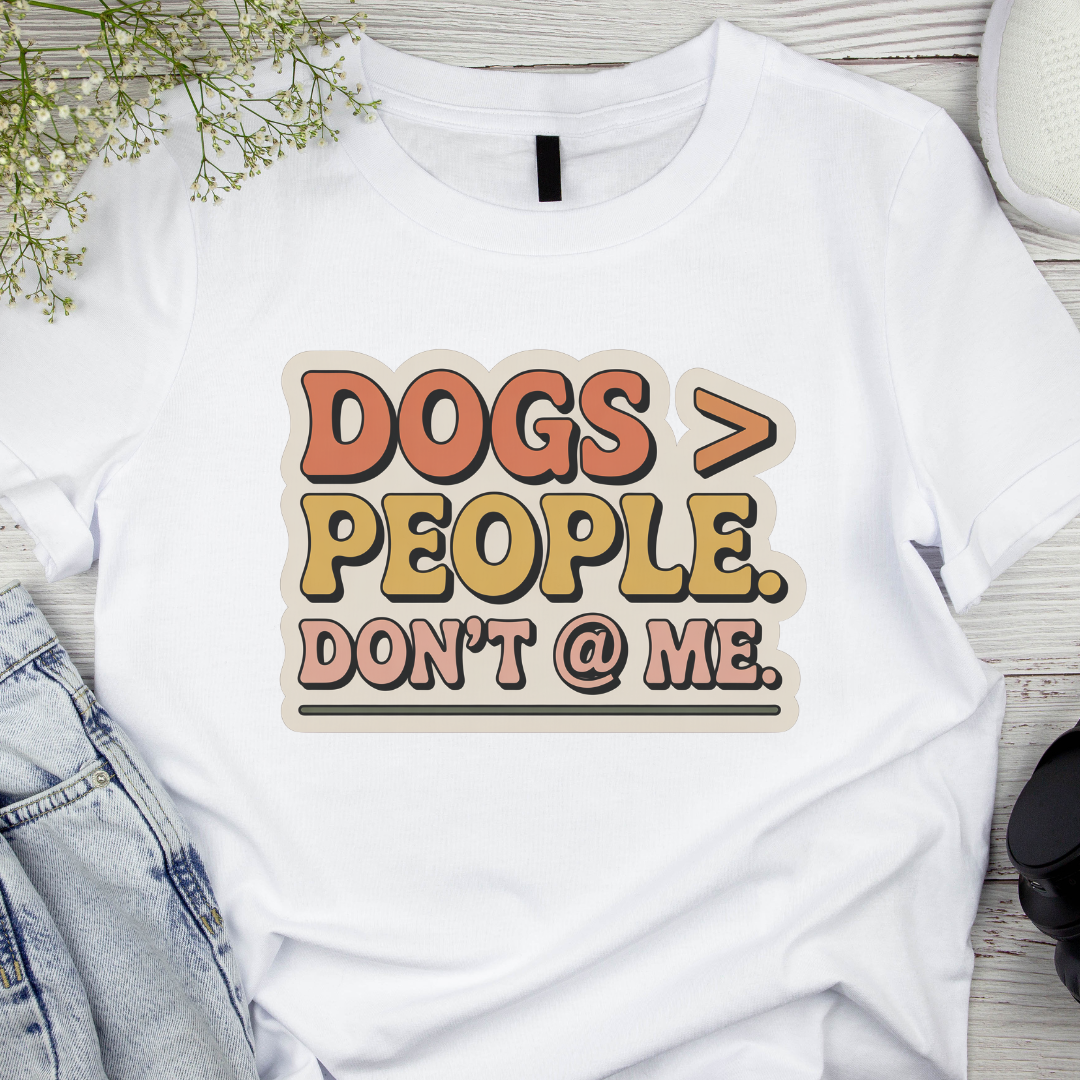 White t-shirt with 'DOGS > PEOPLE. DON'T @ ME.' text on a wooden surface with denim jeans and flowers.