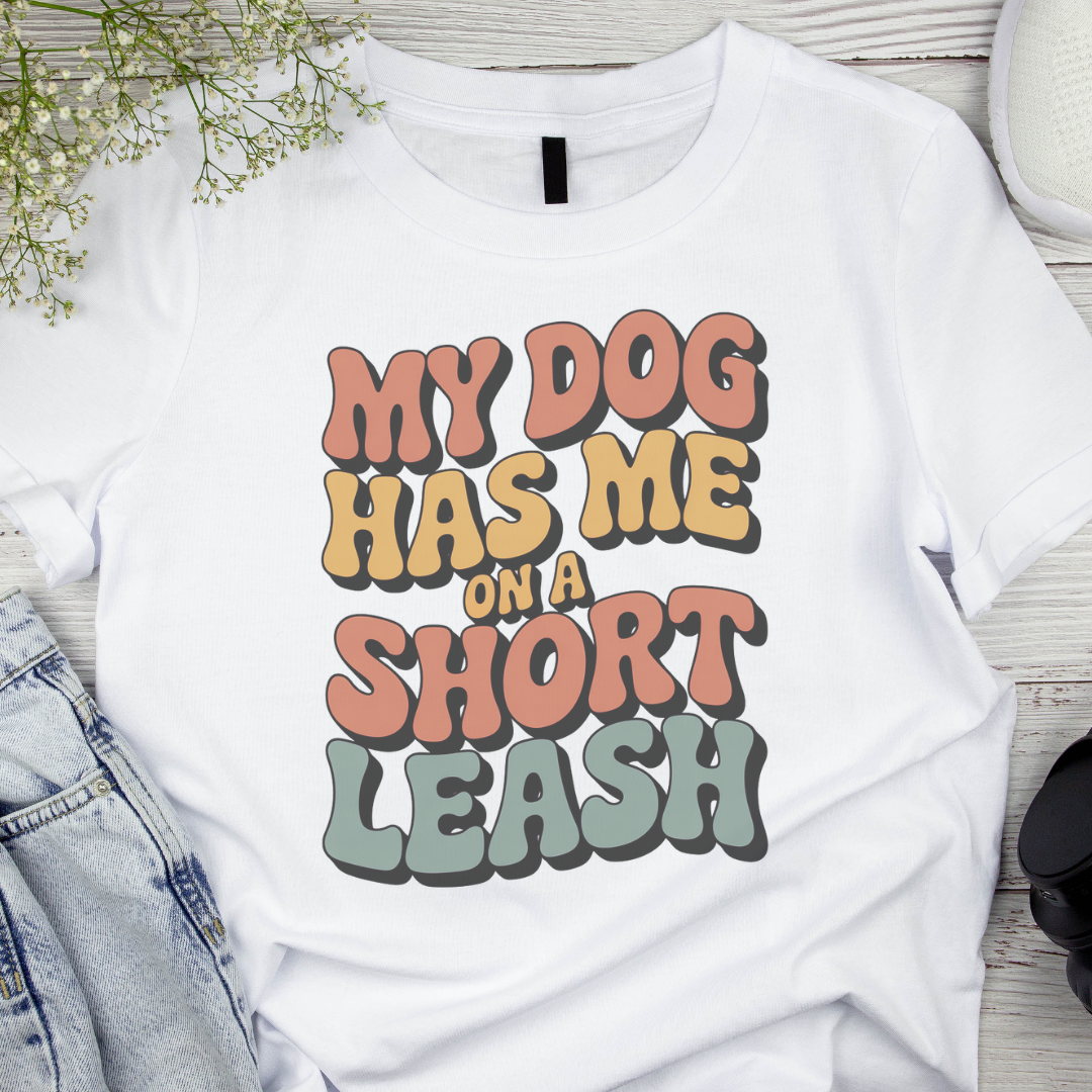 White t-shirt with colorful text 'My dog has me on a short leash' on a wooden surface.