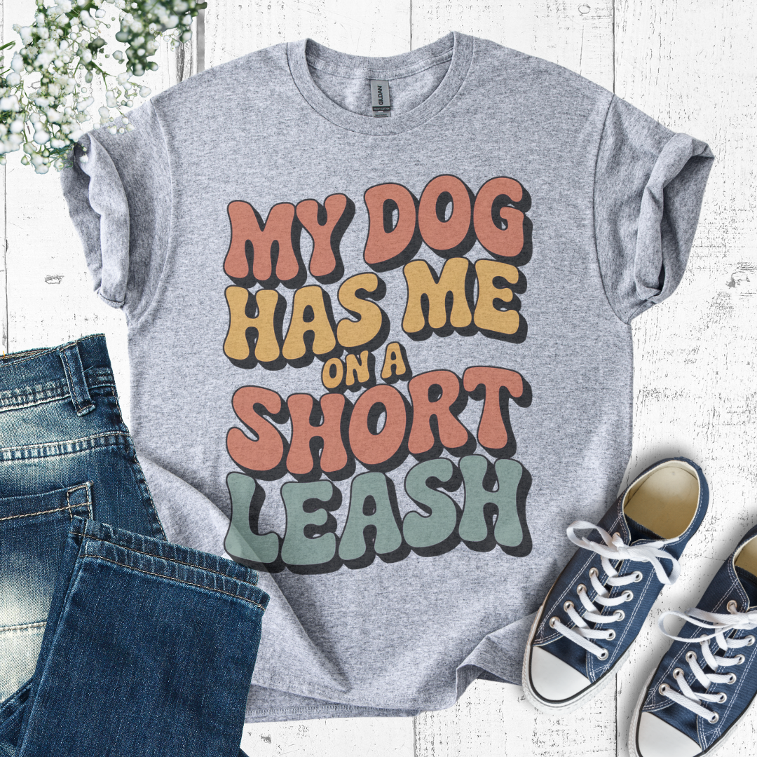 Gray t-shirt with colorful text 'My dog has me on a short leash' on a white background with jeans and sneakers.