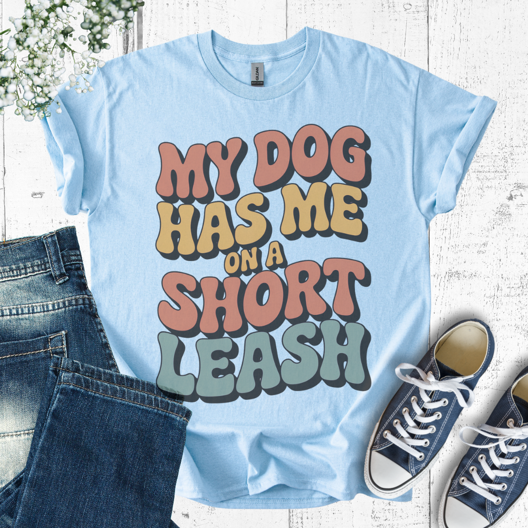 Light blue t-shirt with colorful text 'My dog has me on a short leash' on a wooden surface with jeans and sneakers.