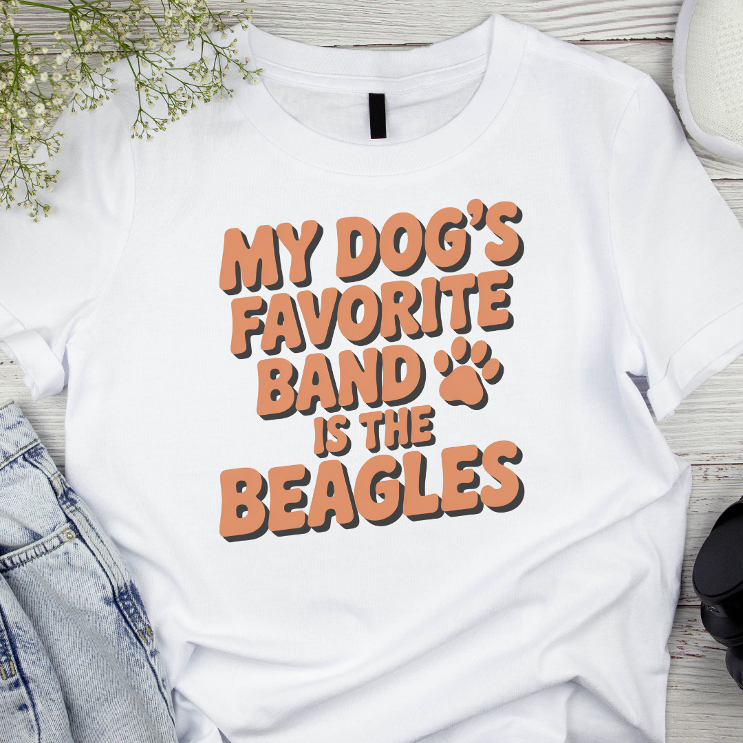 White t-shirt with 'My dog's favorite band is the beagles' text on a wooden surface.