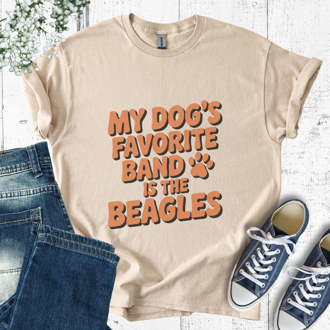 Beige t-shirt with 'My dog's favorite band is the beagles' text, paired with jeans and sneakers on a light wooden surface.