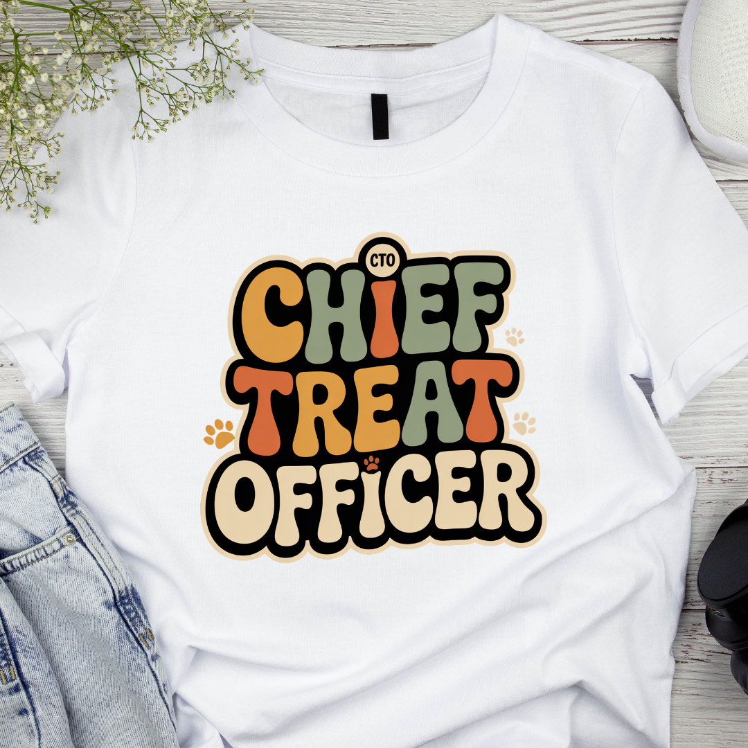 White t-shirt with 'Chief Treat Officer' text on a wooden surface with denim jeans and a plant.