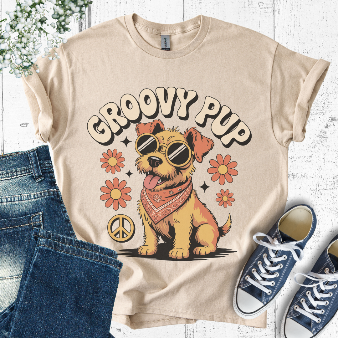 Beige t-shirt with 'Groovy Pup' design featuring a dog, sunglasses, and flowers, paired with jeans and sneakers on a white wooden surface.