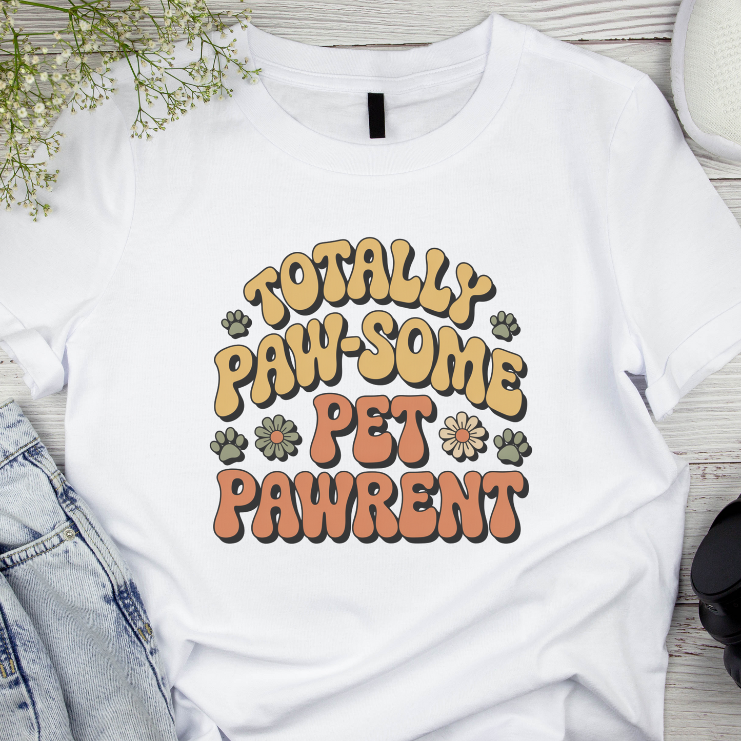 White t-shirt with 'Totally Paw-some Pet Pawrent' text on a wooden surface with denim jeans and flowers.