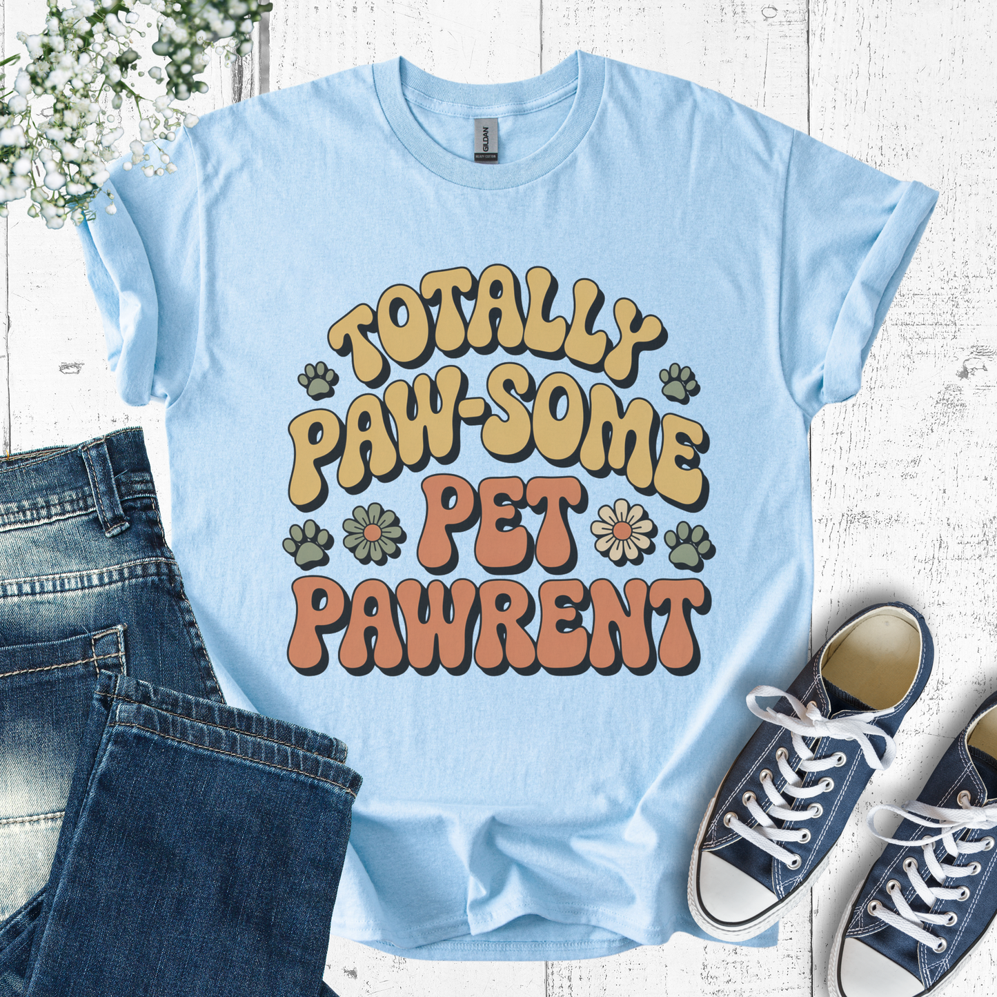 Light blue t-shirt with 'Totally Paw-some Pet Pawrent' text, paired with jeans and sneakers on a wooden surface.