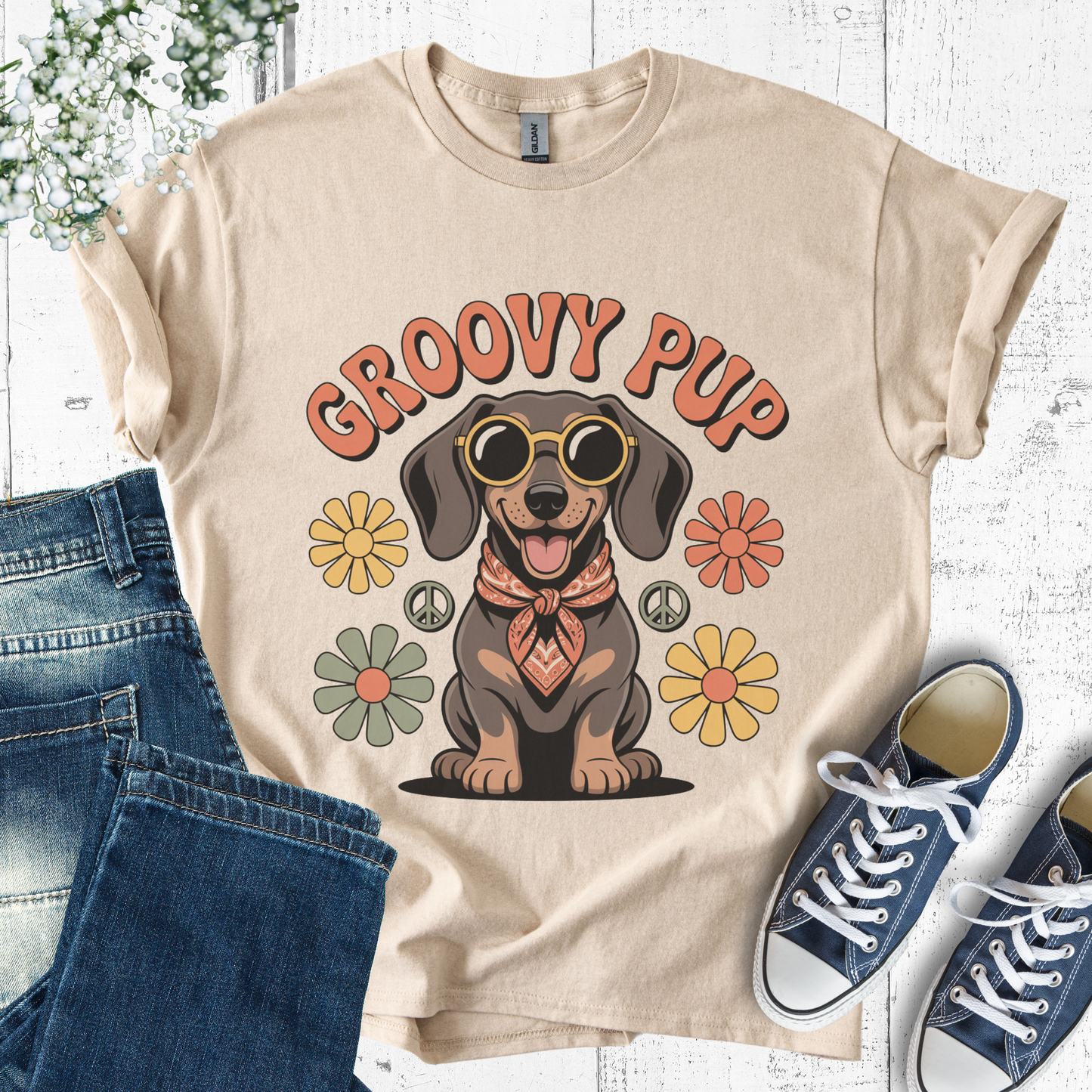 Beige t-shirt with 'Groovy Pup' design featuring a dog, flowers, and peace signs, paired with jeans and sneakers on a white wooden surface.