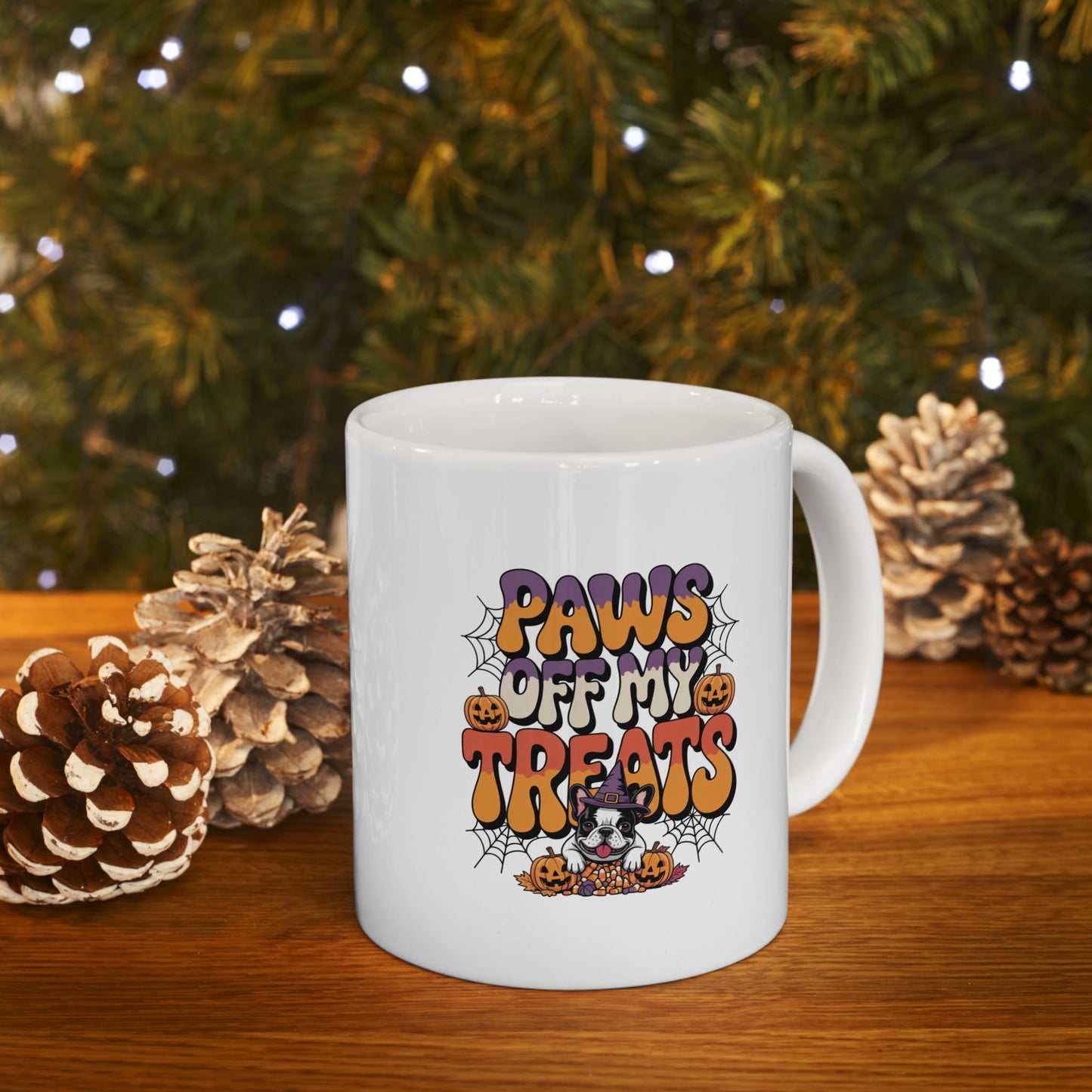 White mug with Halloween-themed design featuring 'Paws Off My Treats' text, placed on a wooden surface with pinecones and a Christmas tree in the background.