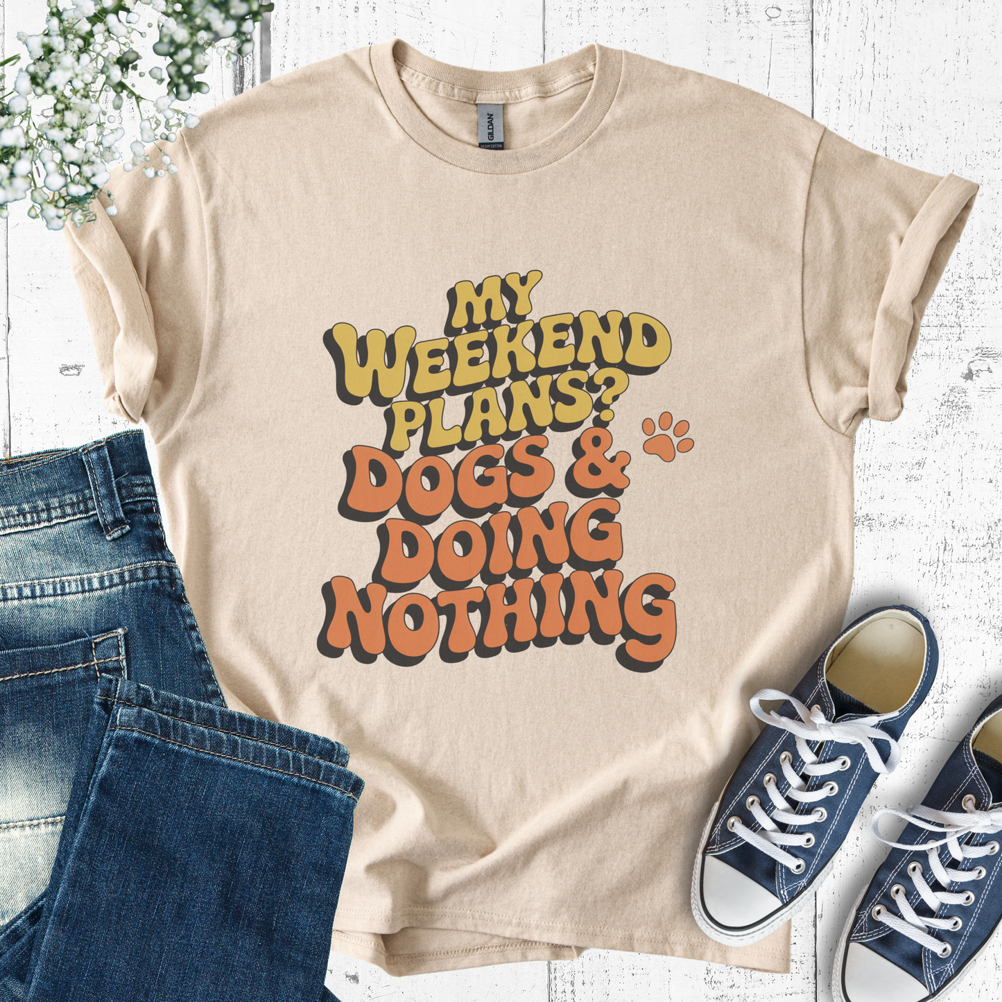 Beige t-shirt with 'My weekend plans? Dogs & doing nothing' text, paired with jeans and sneakers on a white wooden surface.