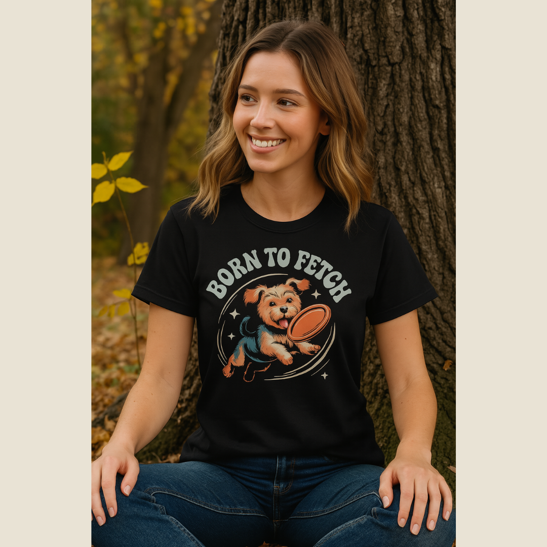 Woman wearing a black t-shirt with 'Born to Fetch' graphic design, sitting against a tree.