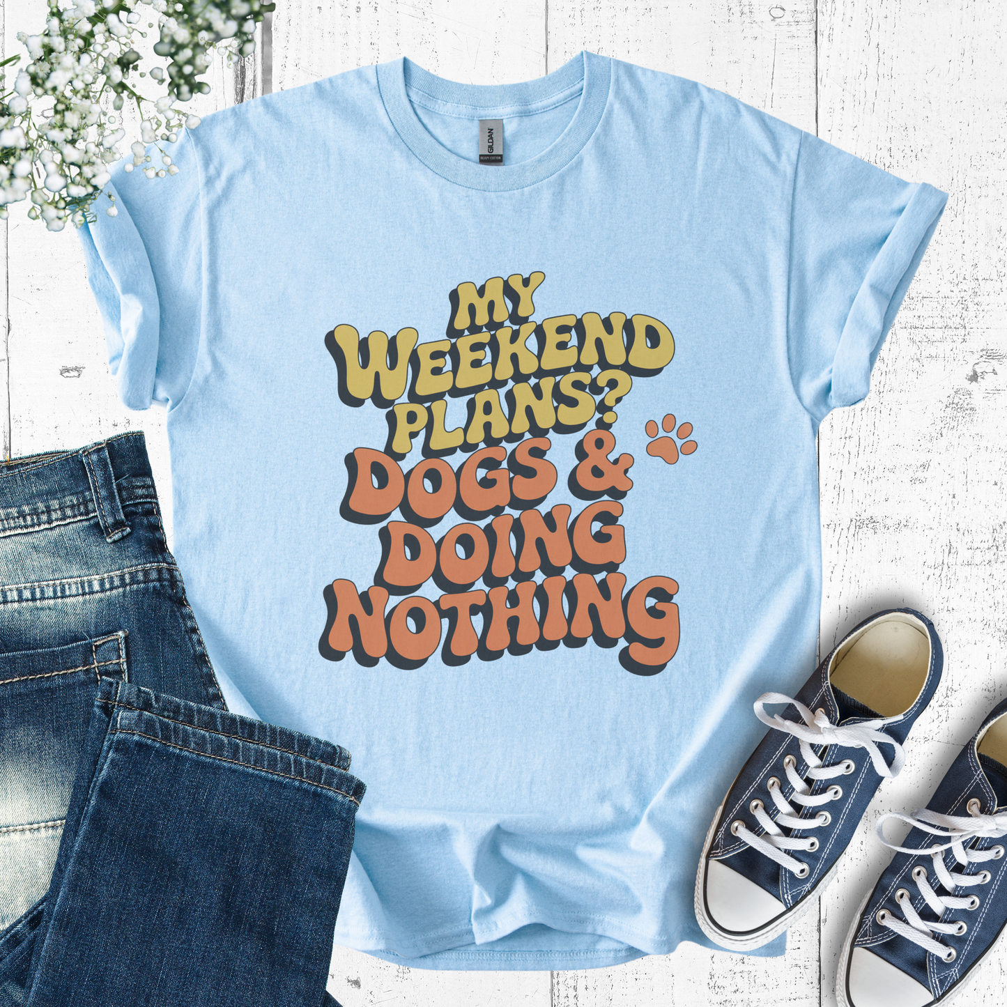 Light blue t-shirt with 'My weekend plans? Dogs & doing nothing' text, paired with jeans and sneakers on a wooden surface.