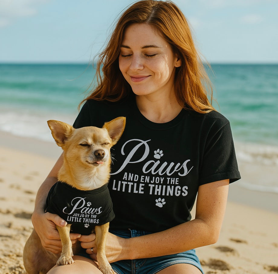 Woman and dog wearing matching 'Paws and enjoy the little things' shirts on a beach.