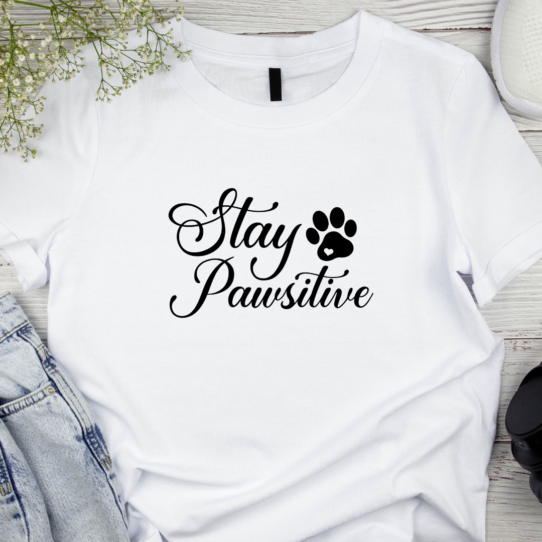 White t-shirt with 'Stay Pawsitive' text and paw print on a wooden surface with denim jeans and flowers.