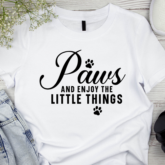 White t-shirt with 'Paws and enjoy the little things' text on a wooden surface.
