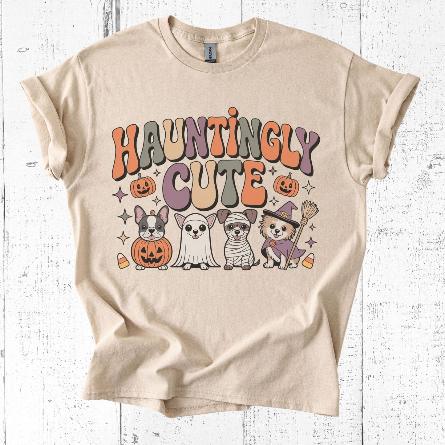 Beige t-shirt with Halloween-themed design featuring cartoon dogs in costumes on a white wooden background.