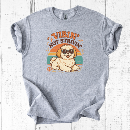 Gray t-shirt with a graphic of a dog wearing sunglasses and text 'Vibin' Not Strivin' on a white wooden background.