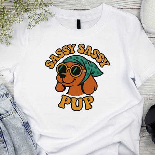 White t-shirt with a graphic of a dog wearing sunglasses and a bandana, with 'Sassy Sassy Pup' text, on a wooden surface.